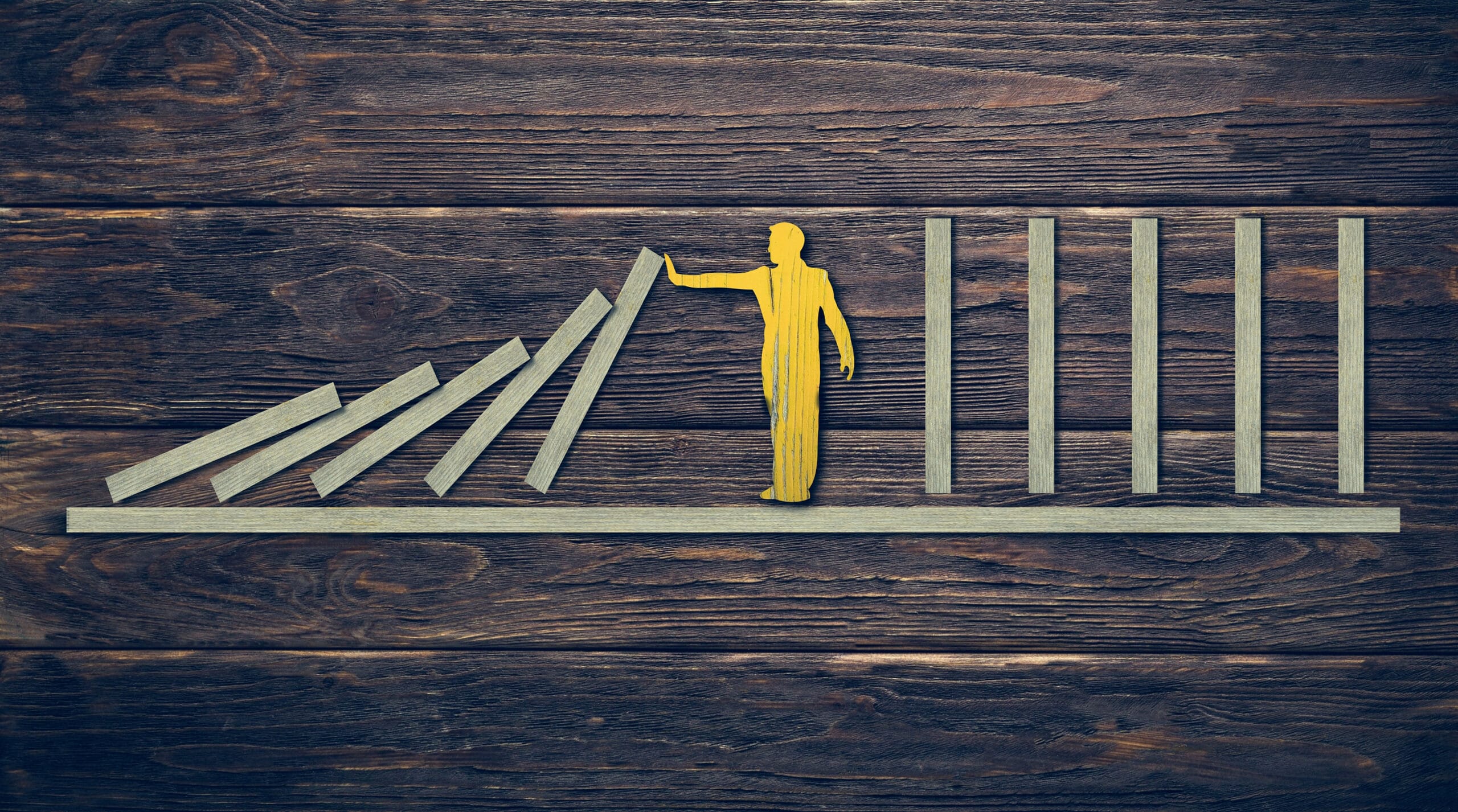 Wooden figure holding up a falling row of domino-like blocks, preventing them from knocking over a line of upright blocks on a wooden surface, symbolizing stability, risk management, or interruption of a chain reaction.